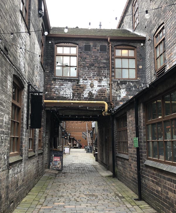 An alleyway between 19th century industrial buildings. The buildings are connected at first floor level, above the alley. Today, the buildings make an attractive scene, with fairy lights.