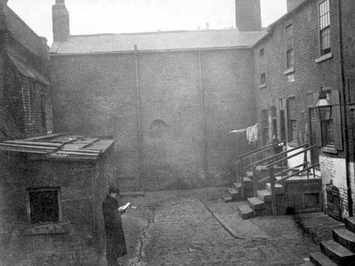 Black and white photograph dated 1901 showing an old yard with two storey houses on each side and steps leading to three of the houses.  A man is standing outside a brick-built lean-to building on the left of the shot.  This may be a privy.